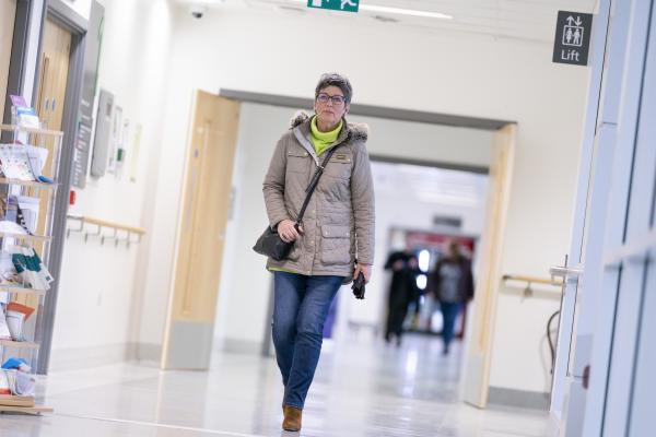 A woman with ovarian cancer wearing a coat and green jumper walking down a hospital corridor