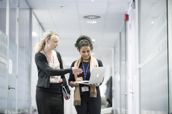 Two women looking at a laptop while walking down a corridor with one woman pointing to the screen