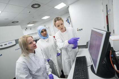 Three female researchers wearing lab coats looking at and pointing to information on a computer screen