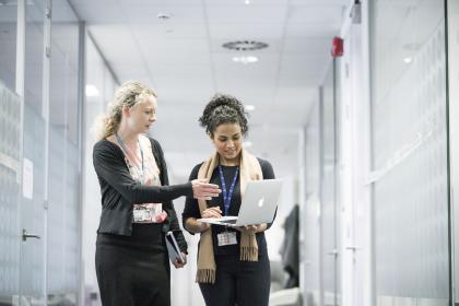 Two women looking at a laptop while walking down a corridor with one woman pointing to the screen