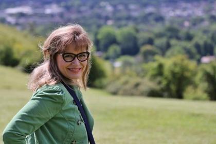 Annie looking toward the camera wearing a green top, scarf and glasses with fields in the background - a woman with ovarian cancer who shared her story with Target Ovarian Cancer