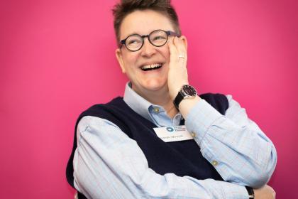 An image of Sarah with her hand touching her face, laughing, with a pink background