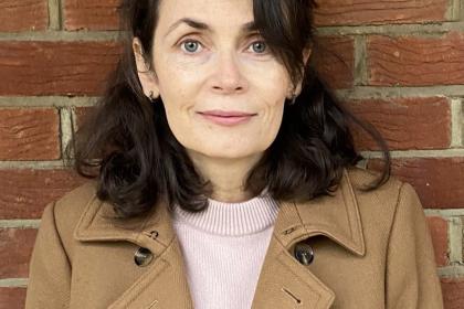 Elaine standing in front of a brick wall wearing a camel colour trench coat and peach top