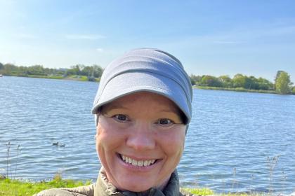 Sarah taking a selfie wearing a cap and grey jacket, she's outside in front of a lake with blue skies behind her