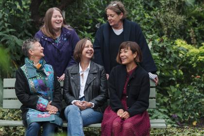 Five women all smiling together in a park. Three are sitting on a bench and two are standing behind them