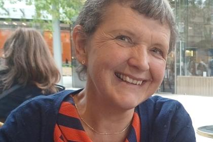 A photo of Jenny smiling. She has short grey hair and is wearing a red patterned top with a navy blue cardigan