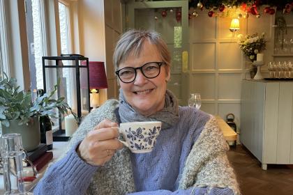 Nicola sitting in a cafe holding a teacup whilst smiling and wearing black glasses and a cosy grey blue jumper