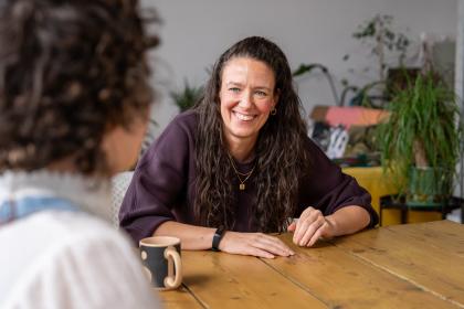 A photo of Lindsay sitting at a table with her hands resting on it smiling. She has long brown curly hair and is wearing a brown top