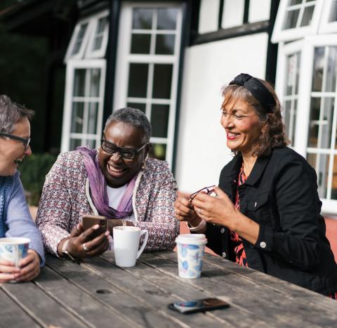 Three women sitting at a bench smiling and laughing together while looking at a phone