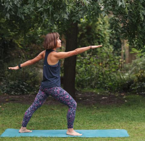 A woman standing in a park on a green yoga mat wearing leggings and a sports top standing in a yoga pose