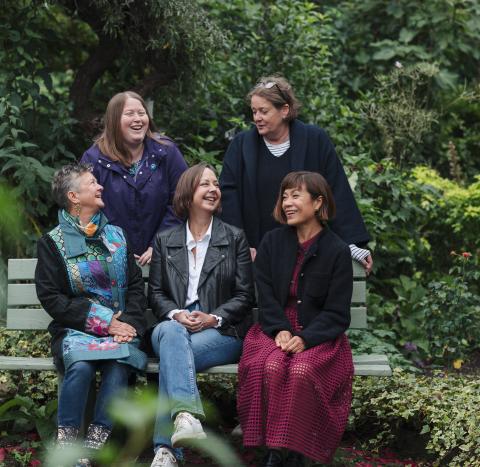 Five women all smiling together in a park. Three are sitting on a bench and two are standing behind them