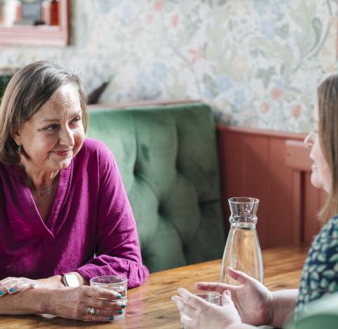 A woman in a pink jumper speaking to another woman across a table.