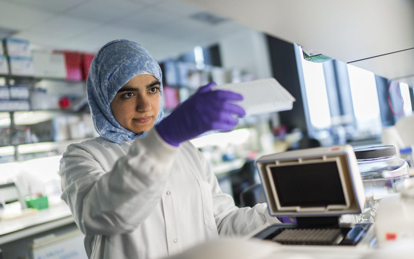 A female ovarian cancer researcher wearing a headscarf, purple gloves and a lab coat using equipment