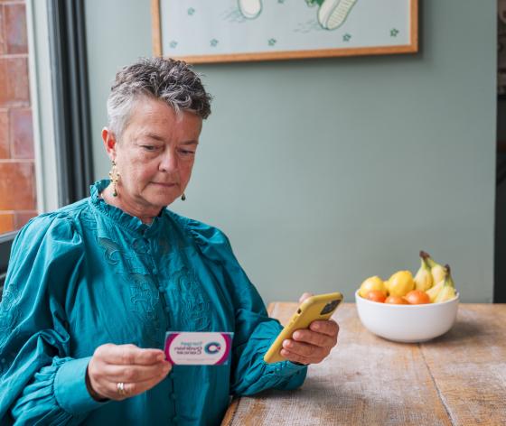 A woman with short grey hair sitting at a table holding her smart phone while looking at a Target Ovarian Business Card