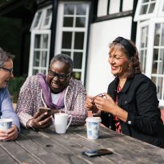 Three women sitting at a bench smiling and laughing together while looking at a phone