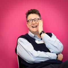 An image of Sarah with her hand touching her face, laughing, with a pink background