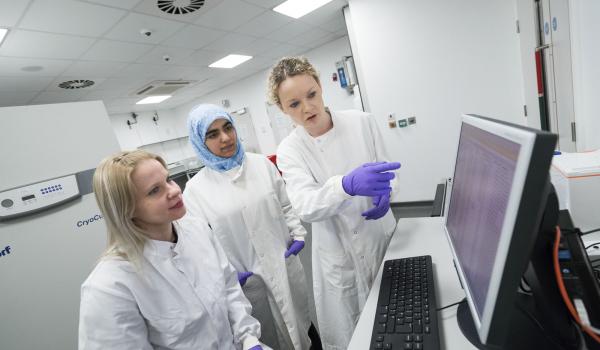Three female researchers wearing lab coats looking at and pointing to information on a computer screen