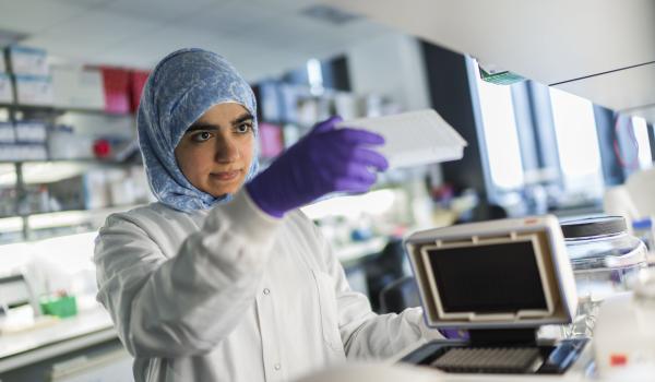 A female ovarian cancer researcher wearing a headscarf, purple gloves and a lab coat using equipment