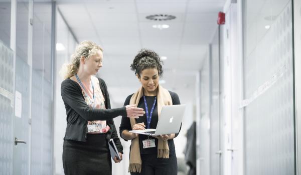Two women looking at a laptop while walking down a corridor with one woman pointing to the screen