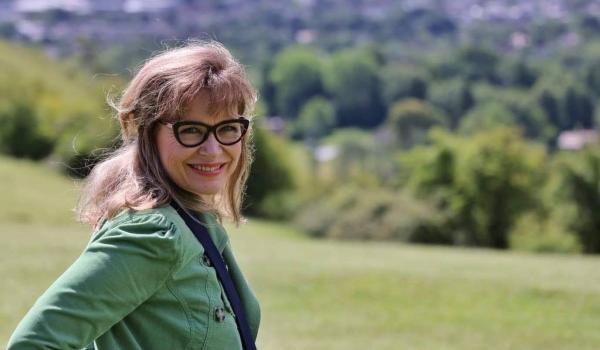 Annie looking toward the camera wearing a green top, scarf and glasses with fields in the background - a woman with ovarian cancer who shared her story with Target Ovarian Cancer