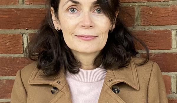 Elaine standing in front of a brick wall wearing a camel colour trench coat and peach top