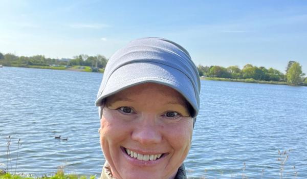Sarah taking a selfie wearing a cap and grey jacket, she's outside in front of a lake with blue skies behind her