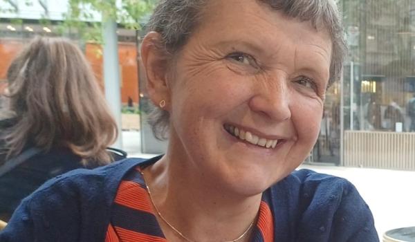 A photo of Jenny smiling. She has short grey hair and is wearing a red patterned top with a navy blue cardigan