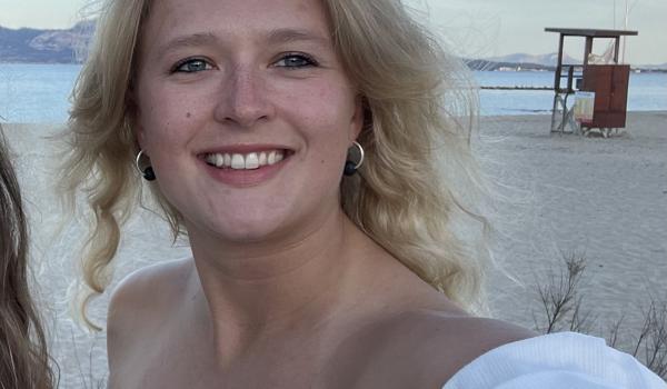 Lotte standing on the beach taking a selfie with wavy blonde hair and wearing a white top