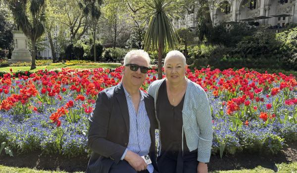Wendy and her husband both standing together and smiling for a photo outside a colourful flower bed