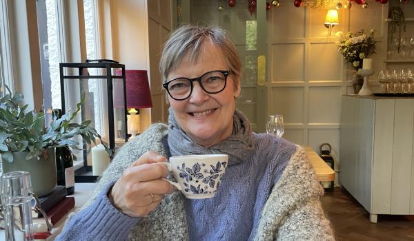 Nicola sitting in a cafe holding a teacup whilst smiling and wearing black glasses and a cosy grey blue jumper