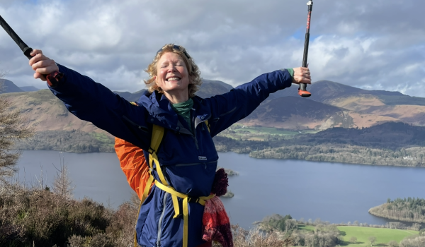 Janet standing on top of a hill with a huge smile, she's holding both of her hands out and up in the air in celebration holding two walking sticks as well