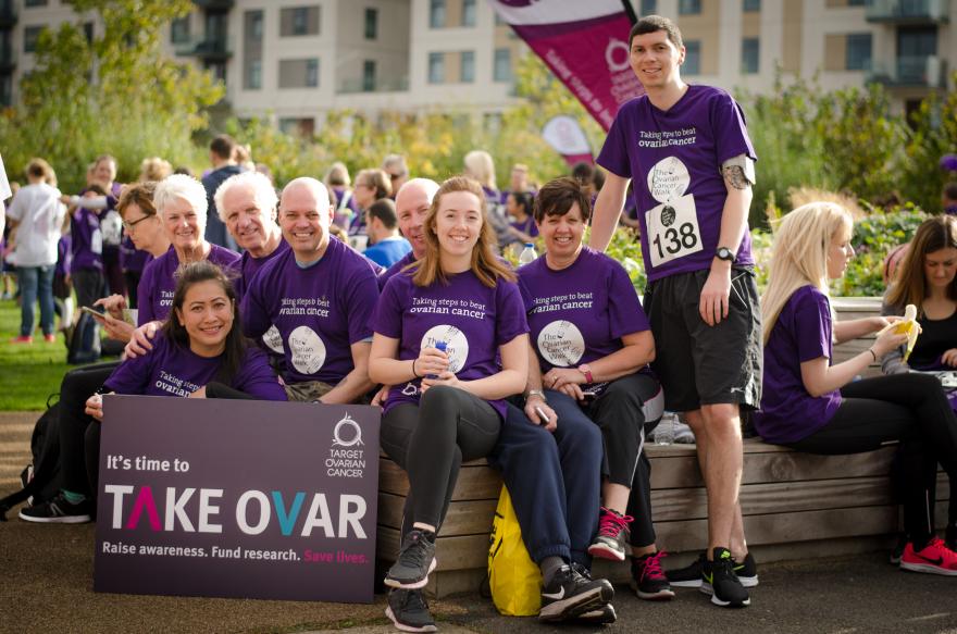 Target Ovarian Cancer supporters sat outside wearing Target Ovarian Cancer Walk Run t-shirts with a TAKE OVAR campaign sign