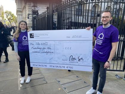 Two people holding a giant cheque for NHS funding wearing purple t-shirts