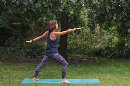 A woman standing in a park on a green yoga mat wearing leggings and a sports top standing in a yoga pose