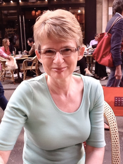 A photo of Julie on holiday smiling towards the camera. She's wearing glasses and a light blue top and has short light brown hair