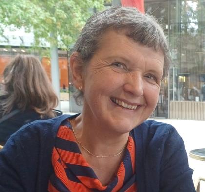 A photo of Jenny smiling. She has short grey hair and is wearing a red patterned top with a navy blue cardigan