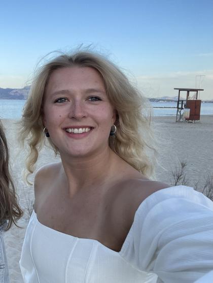 Lotte standing on the beach taking a selfie with wavy blonde hair and wearing a white top