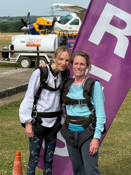 Two women with their arms round each other in skydiving gear pictured on an airfield with a plane in the background