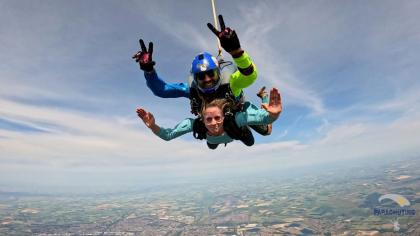 A woman in a blue top doing a tandem skydive with her hands waving to the camera