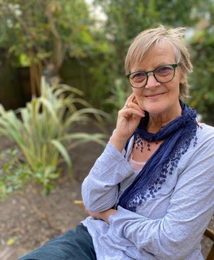 Nicola sitting outside wearing a blue blouse and scarf smiling towards the camera with her hand lightly touching the side of her face