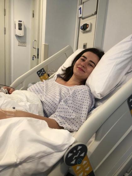 Lucy after her ovarian cancer surgery laying in a hospital bed wearing a hospital gown. She's laying against a pillow and smiling