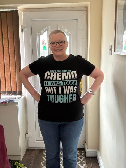 Lesley standing by her front door with her hands on her hips smiling. She is wearing a black tshirt that reads 'My last day of chemotherapy was tough but I am tougher'