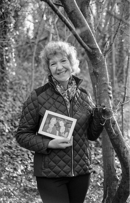Gill standing in the woods smiling whilst holding a photograph of her and her mum