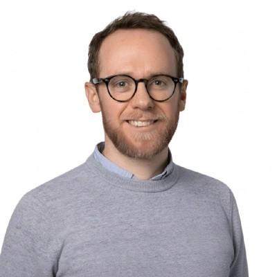 A headshot image of Tim smiling towards the camera. He is wearing a grey jumper and black glasses
