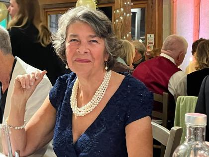 A photo of Georgina sitting at a table smiling. She is wearing a dark coloured dress and a cream necklace