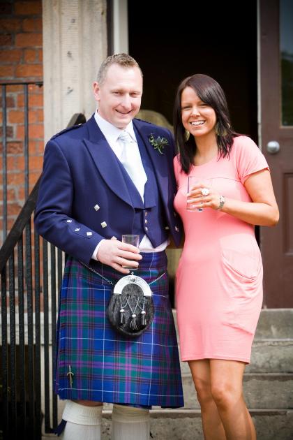 Carl nd Fiona standing smiling together. He is wearing a blue suit with kilt and she is wearing a peach coloured dress