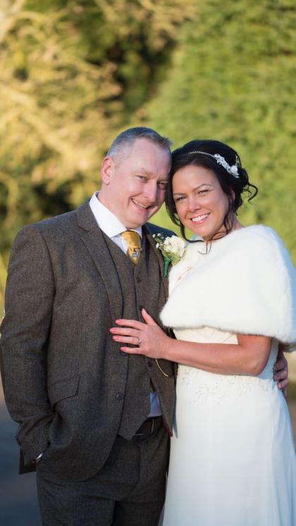  Carl and Fiona on their wedding day. He is wearing a grey suit and she is wearing a white dress with fur trim and has her hand resting on his chest