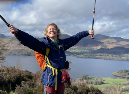 Janet standing on top of a hill with a huge smile, she's holding both of her hands out and up in the air in celebration holding two walking sticks as well