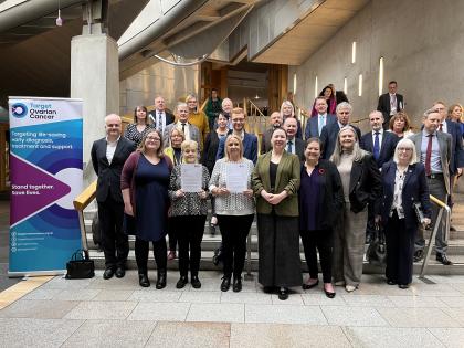 Target Ovarian Cancer campaigners pose with MSPs inside the Scottish Parliament with a letter and beside a Target Ovarian Cancer banner.