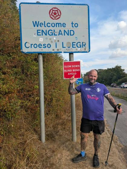 Adam smiles next to a sign that says "Welcome to England" during his seven ultramarathons challenge.
